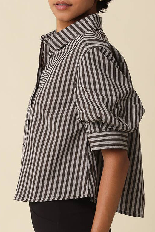 A woman with short curly black hair models a black and white vertically striped short-sleeve button-up shirt, paired with black pants, against a solid beige background.