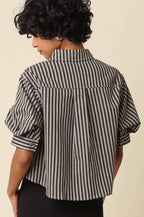 A woman shown from the back wearing a black and white vertically striped short-sleeve blouse with puff sleeves and black pants, against a plain beige studio background.