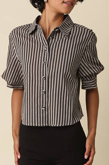 A woman modeling a short-sleeve button-up shirt with black and white vertical stripes, paired with black bike shorts and black Mary Jane shoes, against a solid beige studio background.