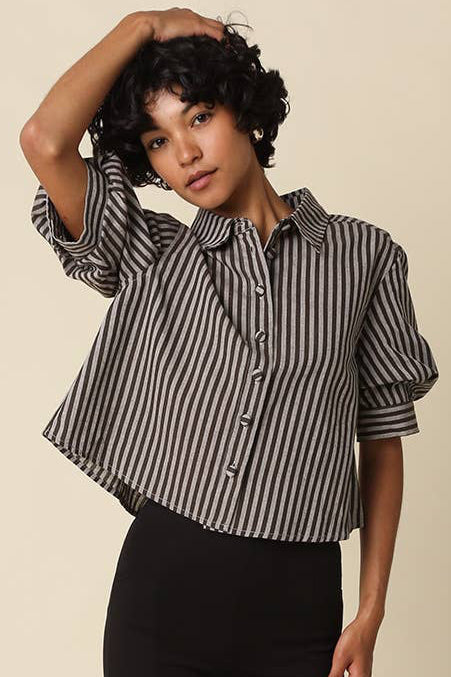 A model wearing a black and white vertically striped short-sleeve button-up shirt, black biker shorts, and black Mary Jane shoes, standing against a solid beige background.