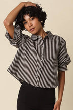 A model wearing a black and white vertically striped short-sleeve button-up shirt, black biker shorts, and black Mary Jane shoes, standing against a solid beige background.