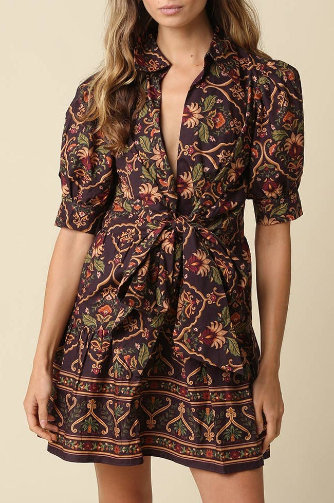 A woman modeling a short button-front shirt dress with a dark brown base and an intricate multicolor floral and paisley print, set against a solid beige background.