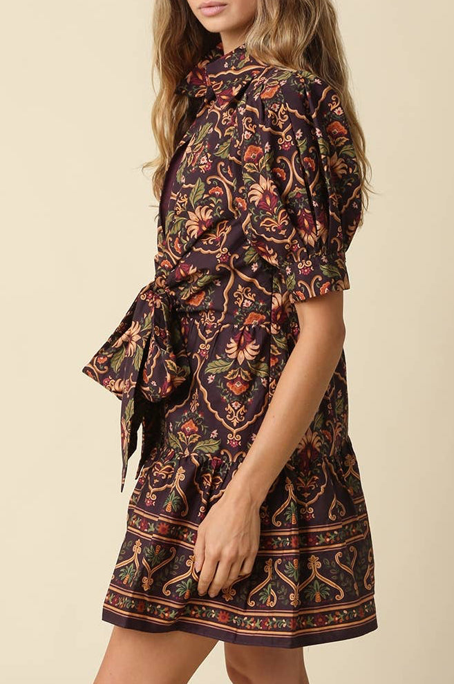 A woman modeling a short-sleeve mini dress with a dark brown base and a colorful floral and paisley print in orange, red, and green, set against a solid beige background.