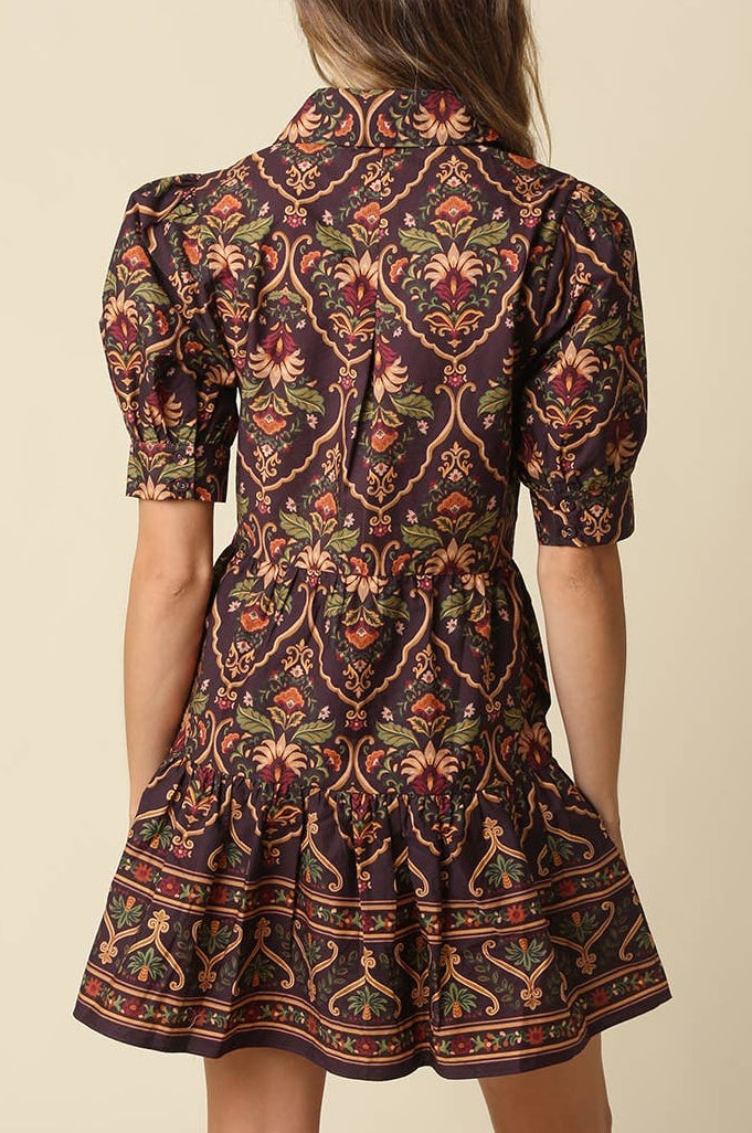 A woman shown from the back wearing a short puff-sleeve dress with a dark brown base and an intricate multicolor floral and scroll pattern in red, green, and gold, set against a solid beige background.