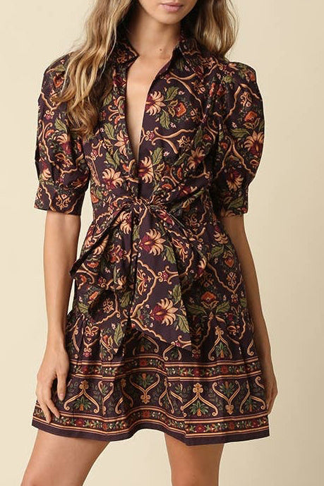 A woman modeling a short puff-sleeve mini dress with a dark brown and black floral pattern and a colorful geometric border, paired with dark brown heeled sandals against a solid beige studio background.