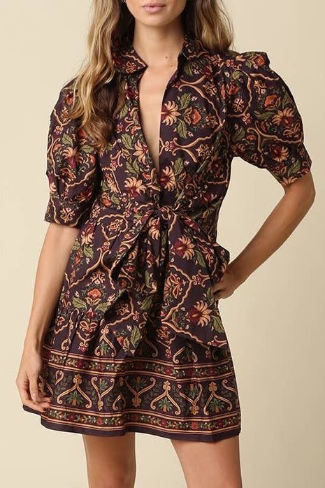 A woman modeling a short puff-sleeve mini dress with a dark brown base and a colorful floral and geometric print, paired with dark brown heeled sandals, against a solid beige studio background.