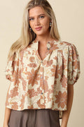 A woman wearing a short-sleeve button-front blouse with a beige base and a rust-orange floral pattern, paired with brown pants, against a solid beige background.