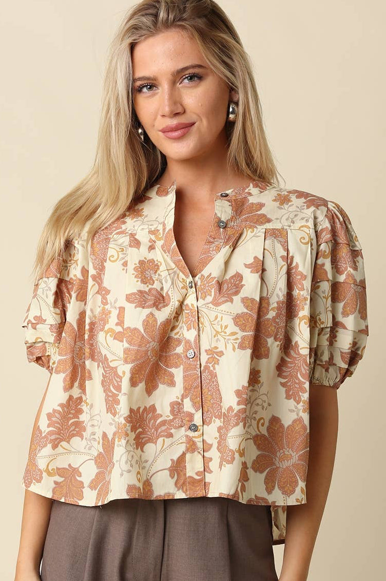 A woman wearing a short-sleeve button-front blouse with a beige base and a rust-orange floral pattern, paired with brown pants, against a solid beige background.