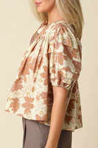 A woman shown in side profile wearing a short-sleeve blouse with a beige and brown floral print, paired with gray pants, against a solid beige background.