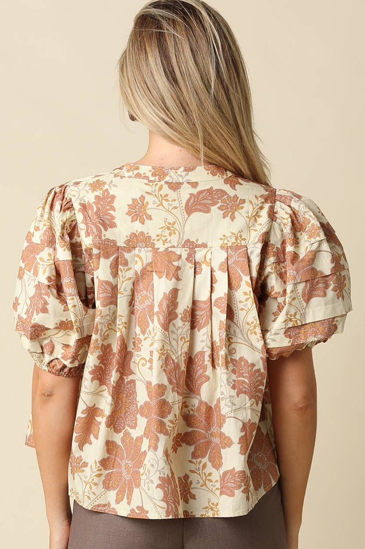 A woman viewed from the back wearing a cream-colored blouse with a dusty rose and brown floral pattern, featuring short puff sleeves and a gathered back. She is paired with brown trousers against a solid beige background.