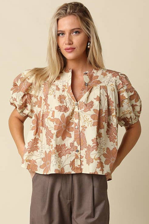 A woman modeling a short-sleeve button-up blouse with a brown and beige floral pattern, paired with wide-leg taupe pants, against a solid beige studio background.