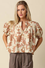 A woman modeling a short-sleeve button-up blouse with a brown and beige floral pattern, paired with wide-leg taupe pants, against a solid beige studio background.