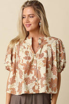A woman modeling a short-sleeve button-up blouse with a beige and brown floral pattern, paired with wide-leg brown trousers, against a solid beige studio background.