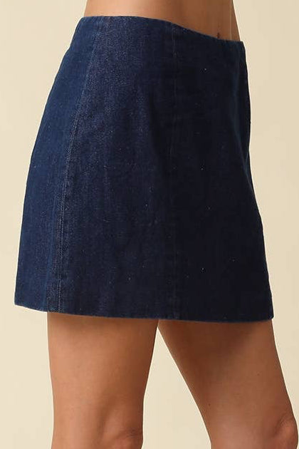 A model wearing a dark blue denim mini skirt paired with black leather knee-high heeled boots, shown from the waist down against a solid beige background.