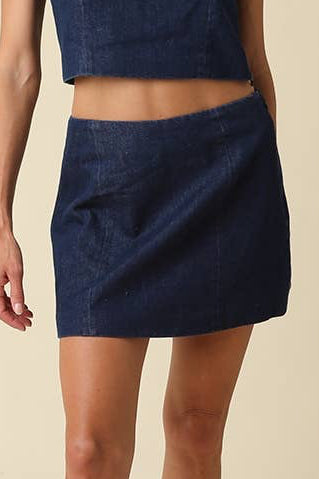 A woman modeling a dark blue denim two-piece set consisting of a sleeveless cropped top and a matching mini skirt, paired with black cowboy boots, against a solid beige studio background.