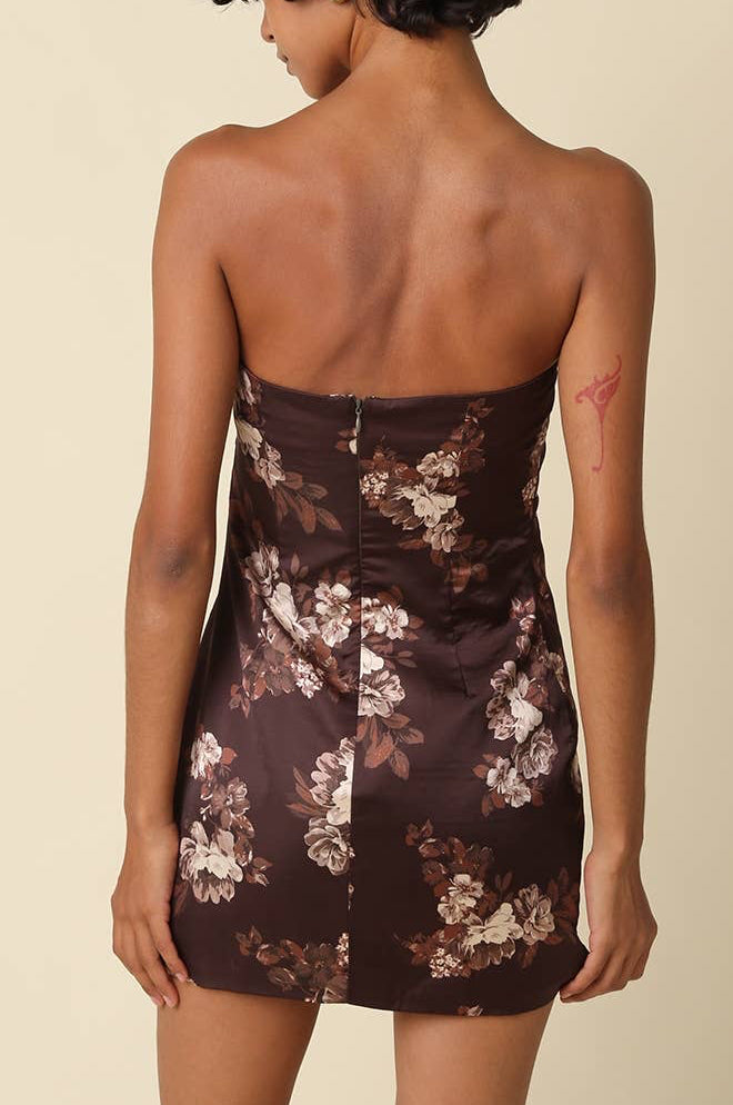 A woman shown from the back wearing a strapless mini dress with a dark brown satin-like base and a floral print in beige, white, and muted pink. The background is a solid, warm beige studio backdrop.