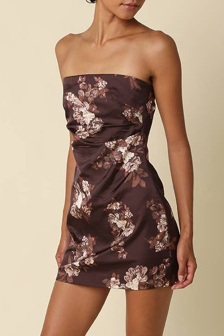 A woman modeling a strapless mini dress with a brown and beige floral print, paired with black heeled sandals, against a solid beige studio background.
