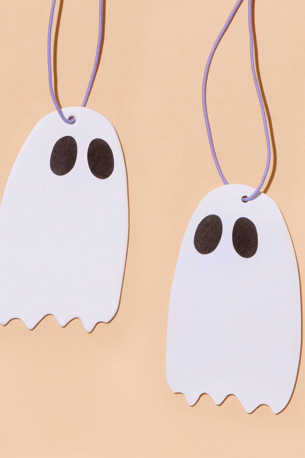 Two white ghost-shaped ornaments with black eyes hanging from thin purple strings against a warm beige background.