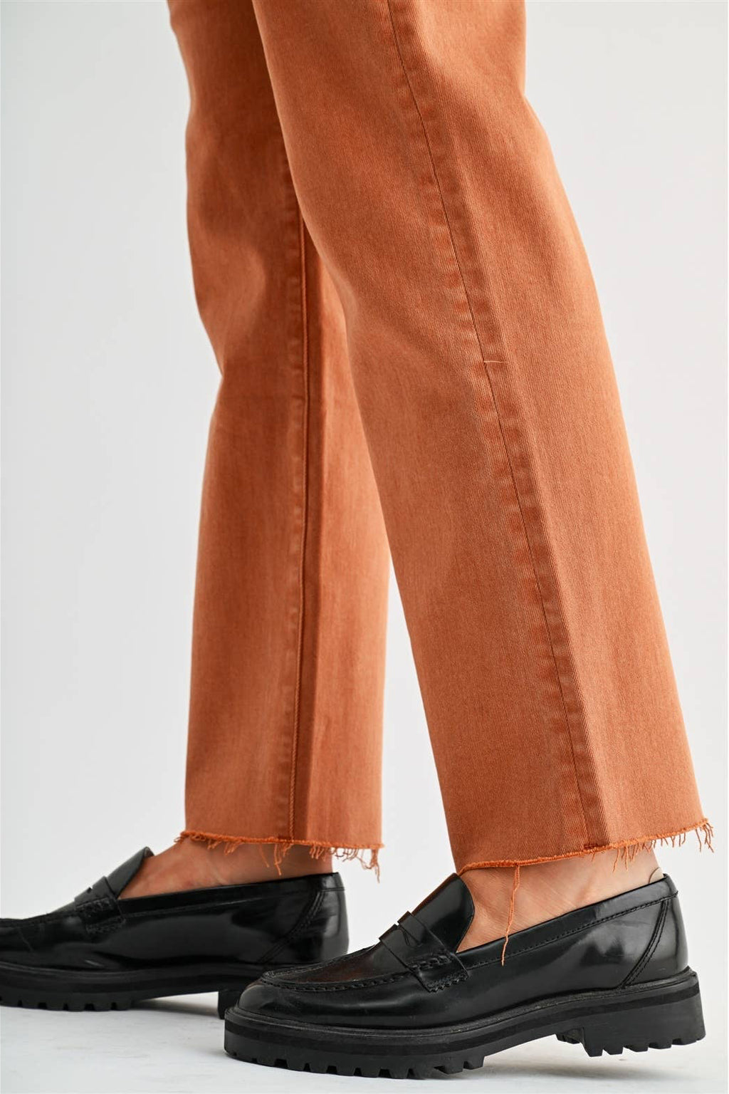 Close-up of a person's legs wearing rust-orange cropped jeans with frayed hems and black leather penny loafers with chunky lug soles, set against a plain light gray studio background.