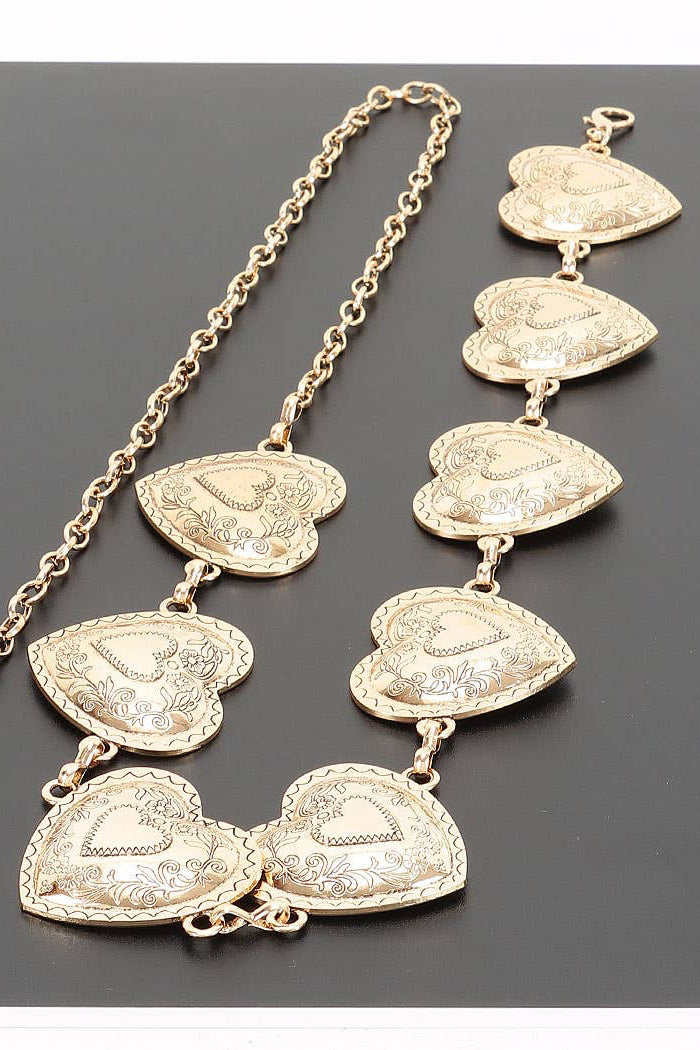 A gold-toned chain necklace featuring multiple textured, heart-shaped pendant links with engraved floral details, displayed on a dark gray background.