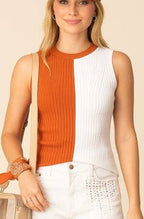 A woman modeling a sleeveless color-block top in orange and white, paired with white denim shorts, a matching orange baseball cap with a white 'T' logo, and white sneakers, holding a small beige bag against a solid beige background.