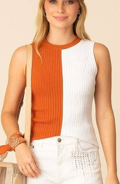 A woman modeling a sleeveless color-block top in orange and white, paired with white denim shorts, a matching orange baseball cap with a white 'T' logo, and white sneakers, holding a small beige bag against a solid beige background.