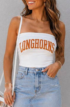A woman modeling a white strapless tank top with 'LONGHORNS' printed in orange across the chest, paired with a light blue denim mini skirt and white cowboy boots, standing against a gray wall.