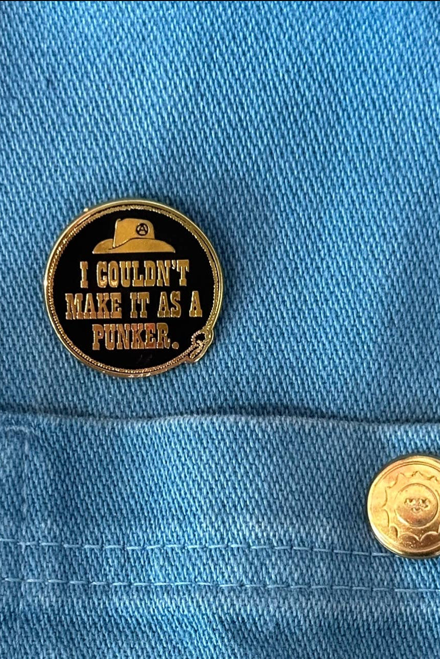 A round gold-tone enamel pin with a black center and text reading 'I COULDN'T MAKE IT AS A PUNKER.' is pinned to a blue denim jacket pocket, next to a matching blue denim snap button.