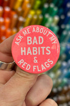 Pink button with text 'Ask me about my bad habits & red flags' held by a hand, with colorful pencils in the background.