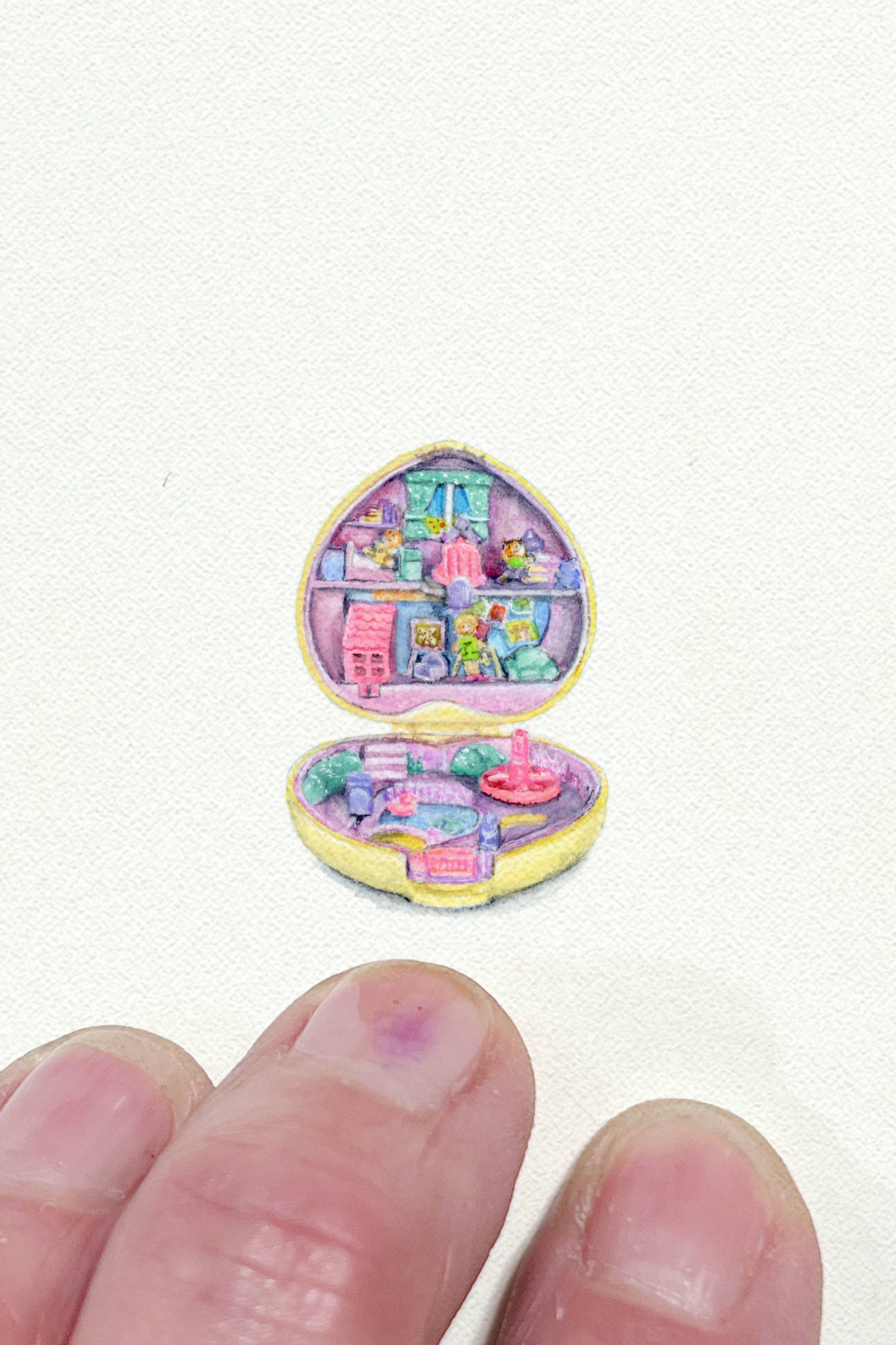 A miniature pastel drawing of an open heart-shaped dollhouse or compact with tiny colorful rooms and furniture, shown on a white textured background with a person's fingers at the bottom for scale.