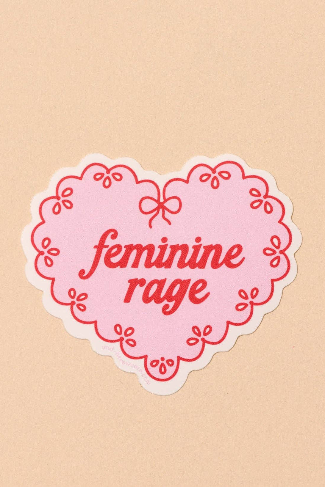 Heart-shaped sticker with 'feminine rage' text on a beige background