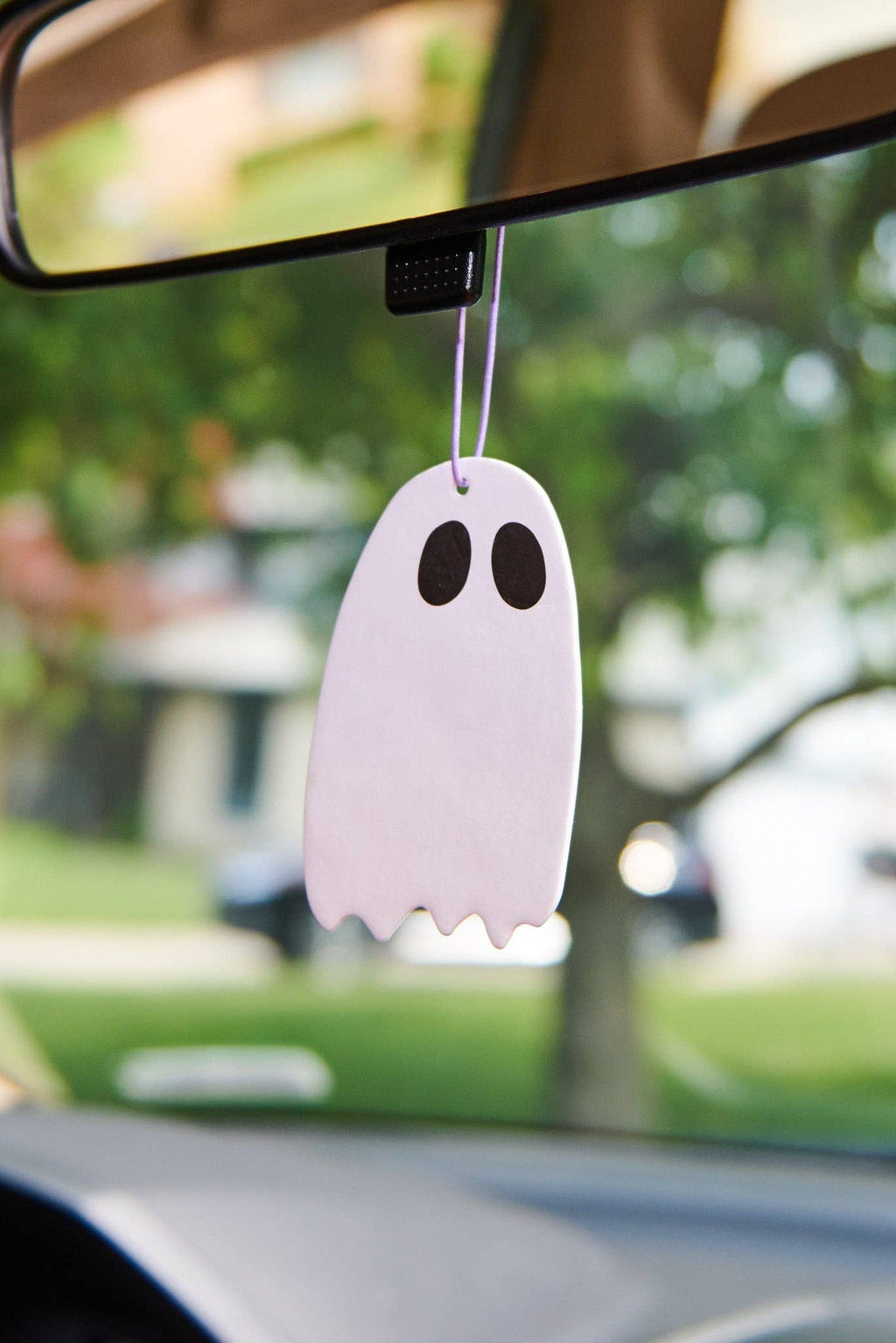 A white ghost-shaped car air freshener or hanging ornament with black eyes hangs from a rear-view mirror inside a car, with a blurred outdoor background visible through the windshield.