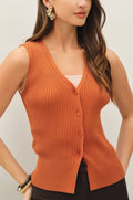A woman wearing a sleeveless ribbed knit cardigan vest in a warm burnt orange color, styled over a white top and dark pants, against a plain light gray background.