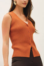 A woman modeling a sleeveless ribbed knit vest in burnt orange paired with dark brown cropped trousers and beige high heels, standing against a plain off-white studio background.