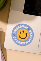 Laptop with a sticker reading 'Probably Overstimulated' on a desk with a notebook and plant.