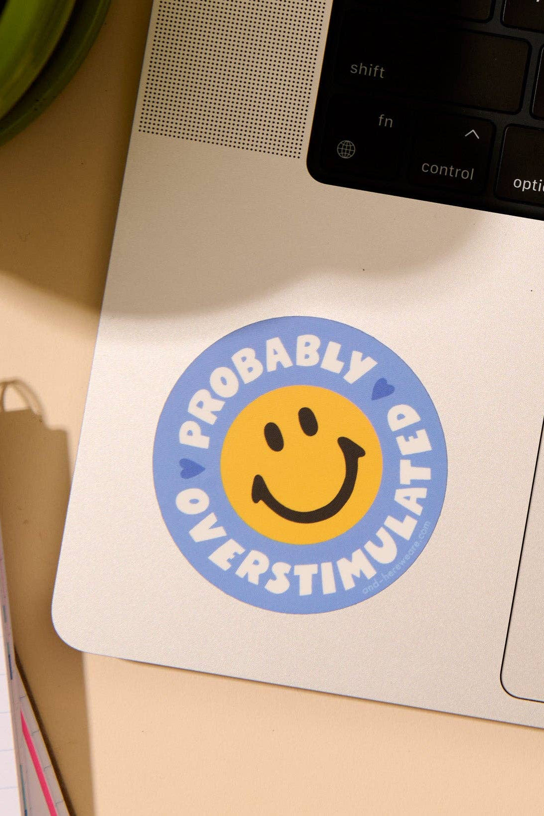 Laptop with a sticker reading 'Probably Overstimulated' on a desk with a notebook and plant.
