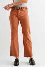 A woman modeling a pair of rust-orange wide-leg jeans with a frayed hem, paired with a patterned belt, a light gray off-the-shoulder t-shirt, and black shoes, against a plain light gray studio background.