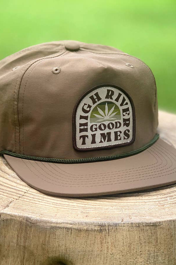 A brown snapback rope hat with 'HIGH RIVER GOOD TIMES' text on a patch, placed on a wooden stump.
