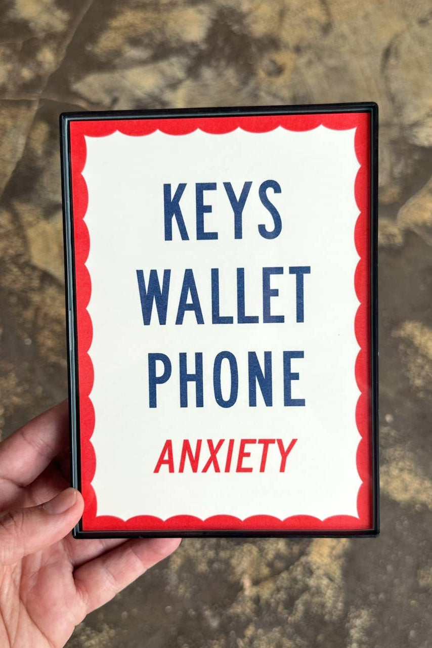 A hand holds a square card or small frame with the words 'KEYS WALLET PHONE ANXIETY' in blue and red text on a white background with a red scalloped border, set against a mottled stone surface.