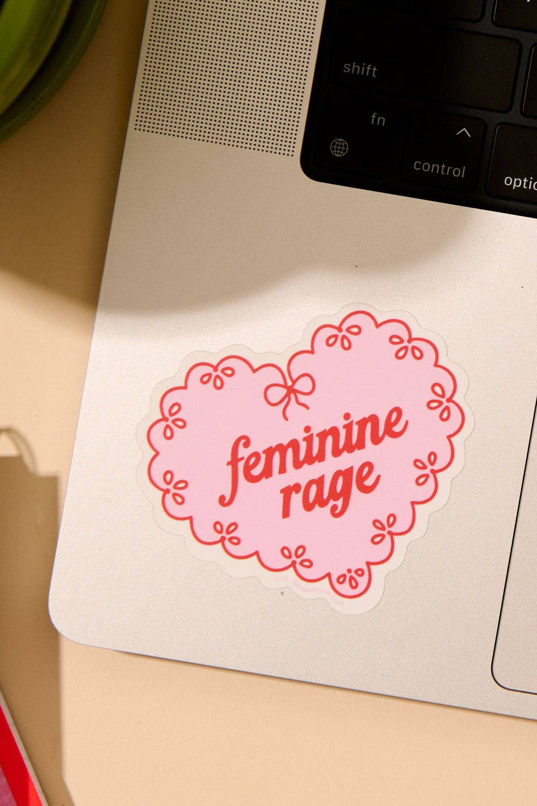 Laptop with a 'feminine rage' sticker on a desk with a notebook and plant.