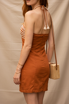 A woman wearing a rust-orange strapless mini dress with a cutout back, walking on a cobblestone street in front of an outdoor cafe. She is carrying a small woven tan bag and wearing brown strappy sandals.