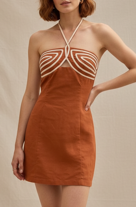 A woman stands on a Parisian cafe sidewalk wearing a rust-orange halter mini dress with white geometric cutout details. She is holding a woven straw handbag and wearing brown sandals, with cafe tables and seated people in the background.