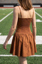 A woman viewed from behind wearing a rust-brown smocked mini dress on a green football field with stadium seating in the background.