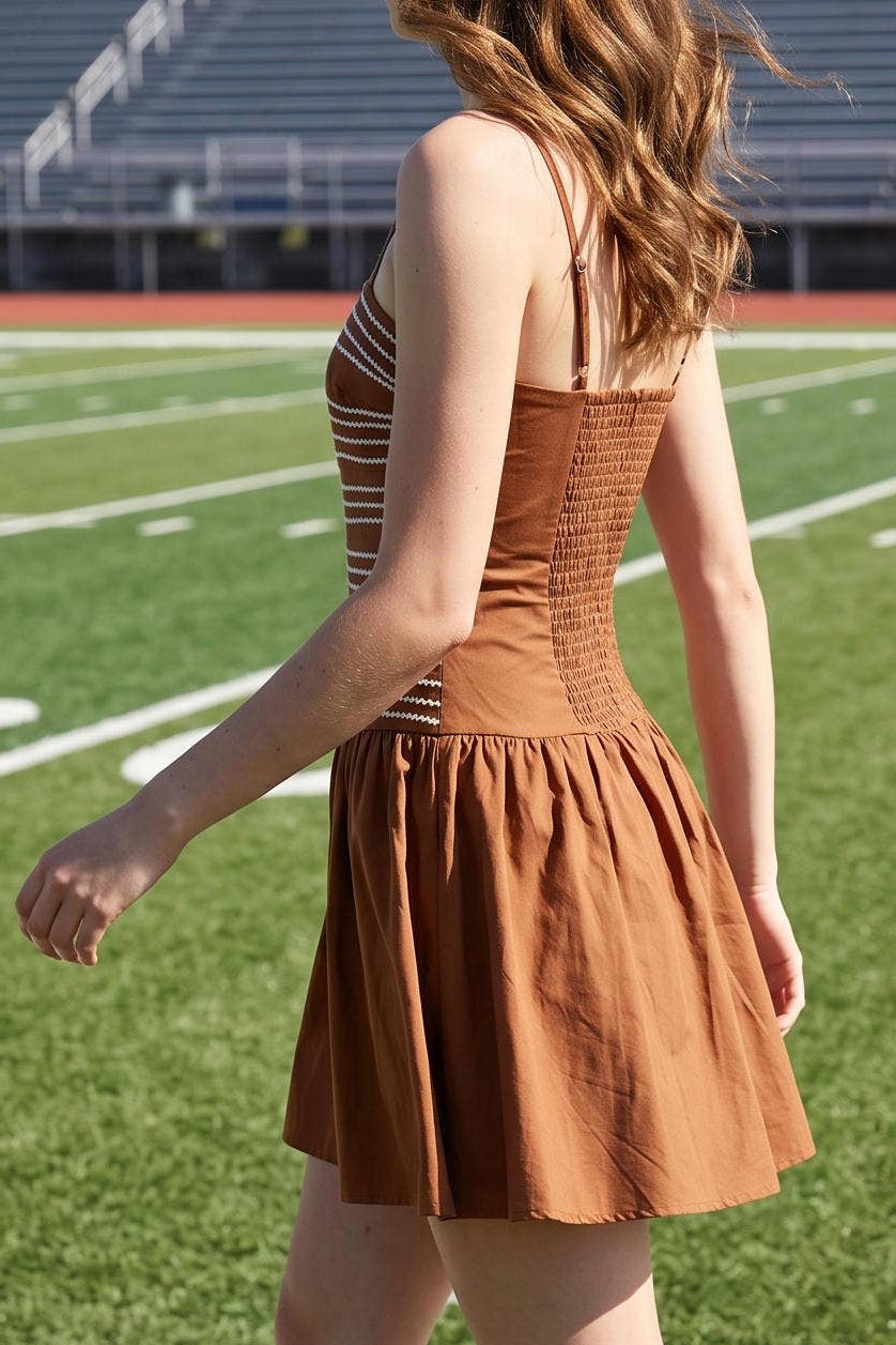 A woman wearing a rust-brown strapless mini dress and white sneakers walks across a white track line on a green football field with stadium bleachers in the background.