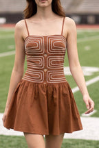 A woman walks on a football field wearing a brown mini dress with a geometric patterned bodice and thin straps, with stadium seating in the background.