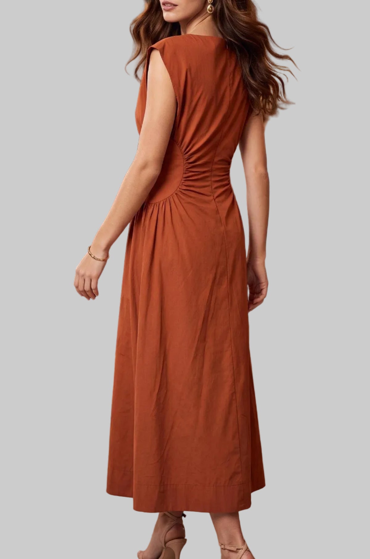 A woman wearing a sleeveless rust-orange midi dress with gathered detailing at the back, standing in a grand ballroom with guests in tuxedos and evening wear.