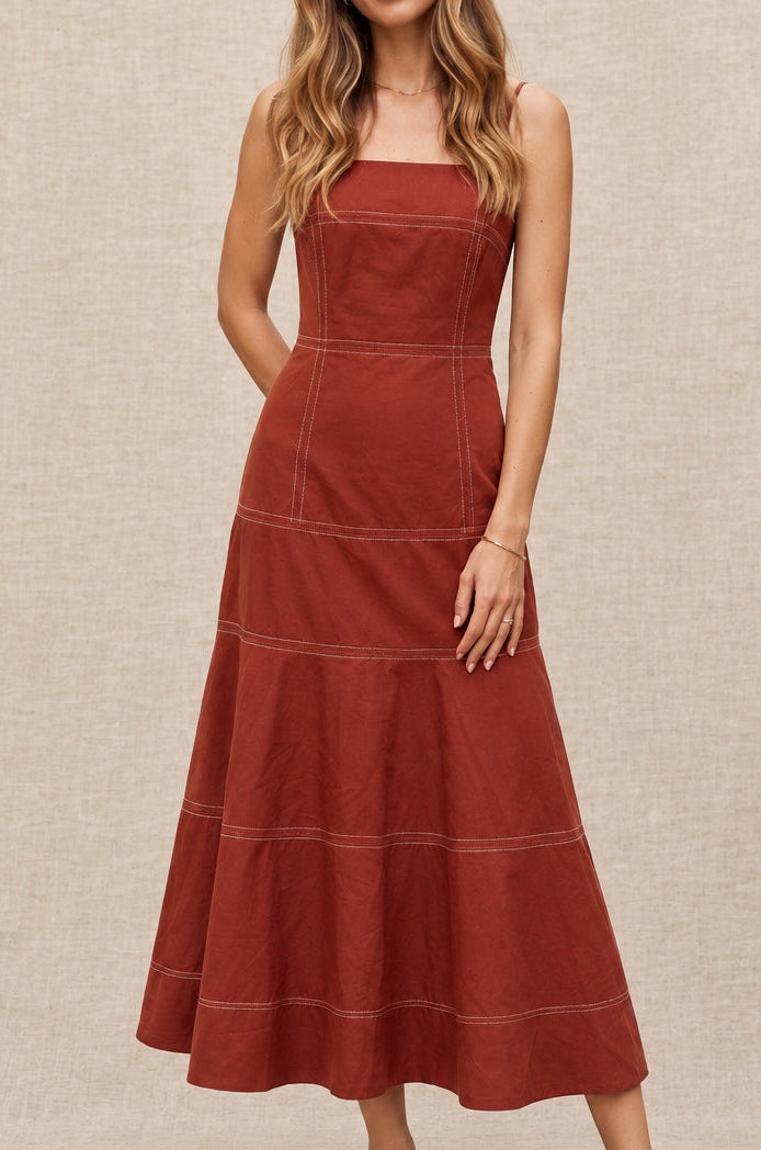 A woman modeling a rust-colored sleeveless midi dress with a structured bodice and tiered skirt, standing on a cobblestone street in a European alleyway.