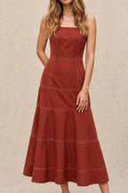 A woman modeling a rust-colored sleeveless midi dress with a structured bodice and tiered skirt, standing on a cobblestone street in a European alleyway.