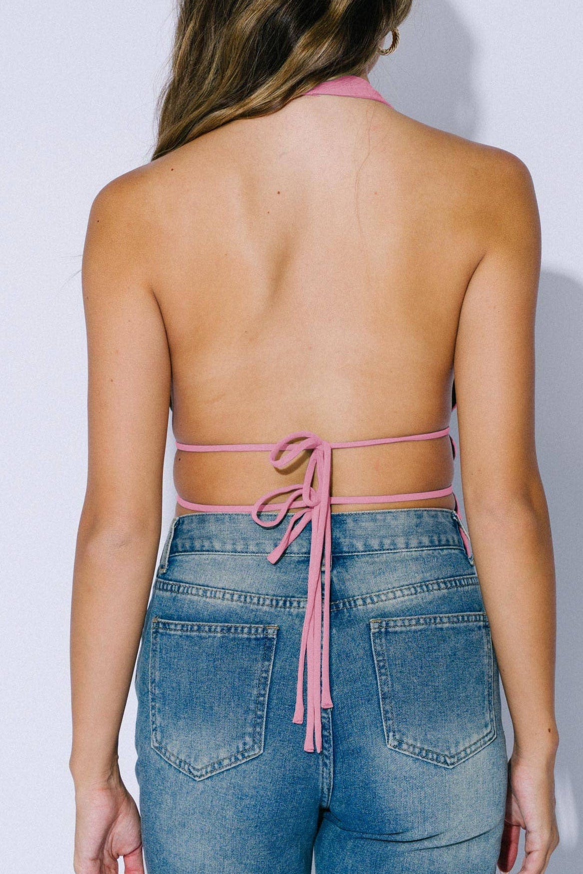 A woman shown from the back wearing a pink halter-style strap top with tie-back straps and blue jeans against a plain light gray studio background.