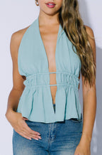 A woman modeling a light blue sleeveless halter top with a deep plunging neckline and a gathered peplum waist, paired with blue jeans against a plain light background.